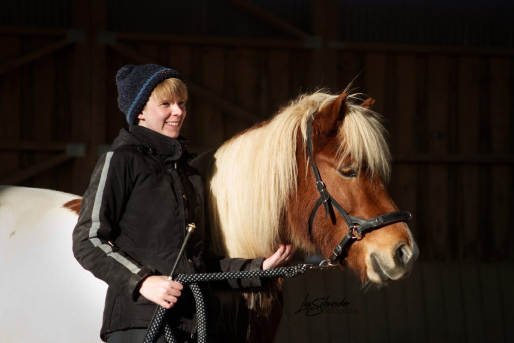 Carina Warnstädt mit Islandstute Eine Frau mit schwarzem Anorak und dunkler Mütze steht lächelnd neben einem braun-weiß geschecktem Islandpony und hält es am Strick fest, der am braunen Halfter befestigt ist.