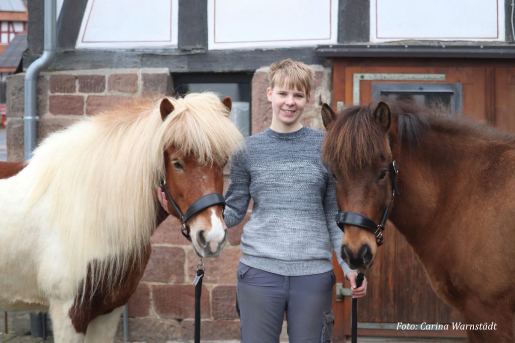 Carina Warnstädt Eine blonde Frau in grauem Pulli und grauer Hose steht lächelnd zwischen einem braun-weiß geschecktem und einem braunen Islandpony.