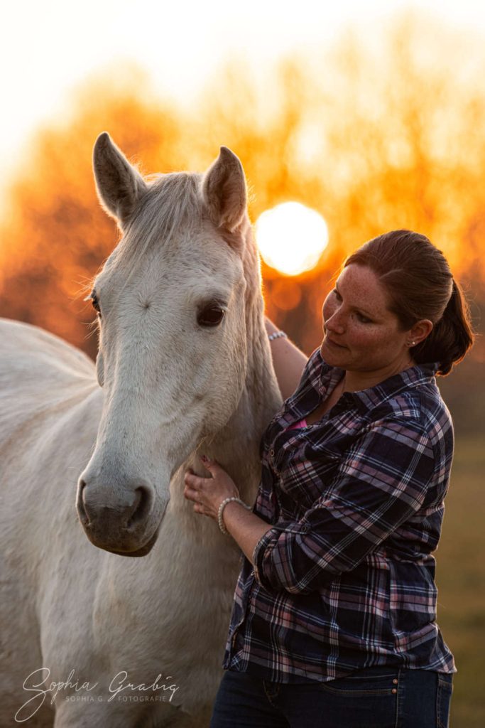 Anna braunhaarige Frau mit karierter Bluse und ein weißes Pferd mit untergehender Sonne und Abendrot im Hintergrund.